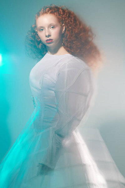Art and fashion. Portrait of a refined fashion model girl with lush red curly hair posing in a white haute couture dress. Studio shot. 