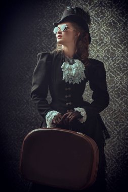 Geleneksel 19. yüzyıl kıyafetleri içinde zarif bir kadının portresi ve şık güneş gözlükleri elinde seyahat çantasıyla klasik bir arka planda poz veriyor. 19. yüzyılın sonları - 20. yüzyıl başları.