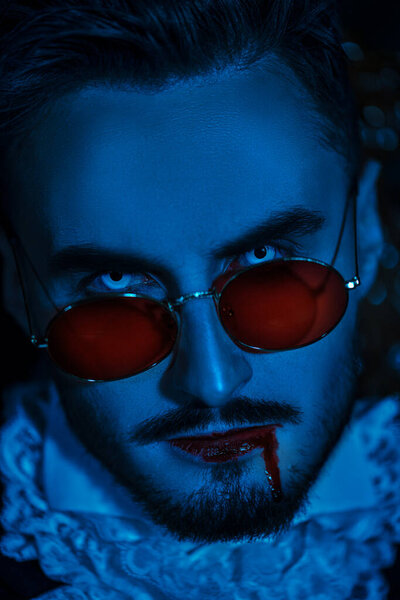 Close up portrait of a handsome blue-eyed vampire man with blood on his lips wearing an elegant 19th century suit and red sunglasses. Halloween. Vampires. 
