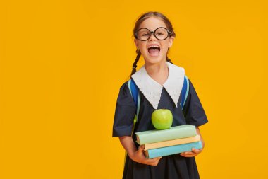 Okul üniformalı, okul çantalı, kitaplı ve elmalı küçük tatlı bir kız sarı arka planda stüdyoda poz veriyor. Eğitim. Metin için yer.