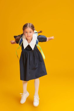 Eğitim. Okul üniformalı ve sarı bir stüdyo geçmişine atlayan okul çantasıyla neşeli küçük bir kızın tam boy portresi. Metin için yer.