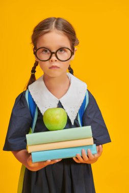 Okul üniformalı, okul çantalı, kitaplı ve elmalı bir kız çocuğu sarı arka planda stüdyoda poz veriyor. Eğitim. 