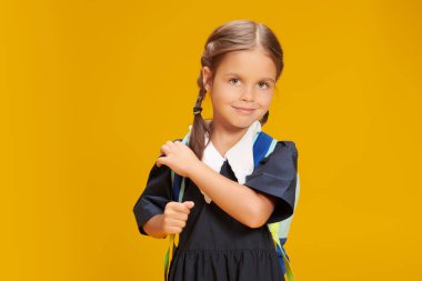 Eğitim. Okul üniformalı ve okul çantasıyla stüdyoda sarı arka planda poz veren küçük tatlı bir kız öğrenci. Metin için yer.