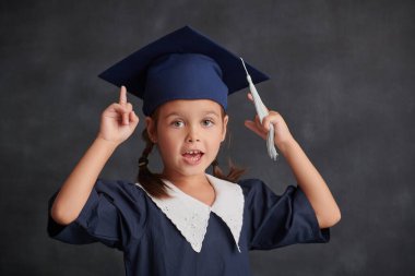 Eğitim. Okul üniformalı ve akademik şapkalı meraklı küçük bir kız siyah bir okul yönetim kurulunun arka planında duruyor ve işaret parmağını kaldırıyor - yeni bir fikir. Metin için yer.