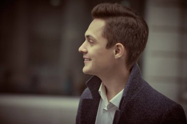 Paltolu yakışıklı, modern bir adam bulutlu bir günde bir şehir caddesinde duruyor ve hafifçe gülümsüyor. İnsanlar ve duygular, yaşam tarzı.