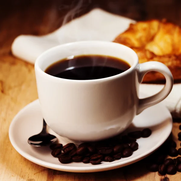 Vit kopp kaffe, croissant och kaffebönor på träbord. ( — Stockfoto