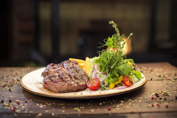 쇠고기 스테이크, 감자, 샐러드 나무 테이블, 벽난로 근처에 맛 있는 저녁 식사 — 스톡 사진