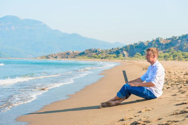 Joven guapo con un ordenador portátil sentado en la playa — Foto de Stock