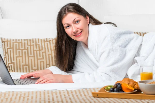 Mooi meisje met een laptop in badjas op het bed — Stockfoto