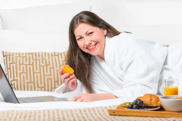 Woman with orange in her hand on the bed in the morning working — Φωτογραφία Αρχείου