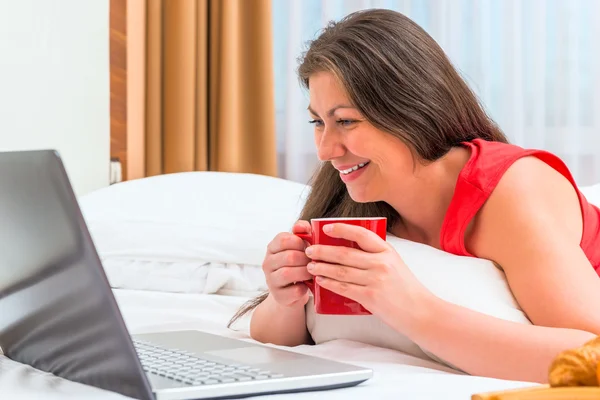 Smiling girl attentively looks at a laptop and drinking tea — Stock Photo, Image