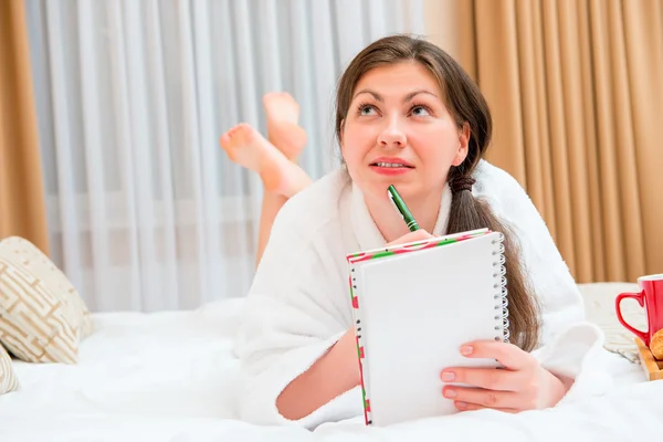 Attractive brunette with a notebook and pen on the bed — Stock Photo, Image