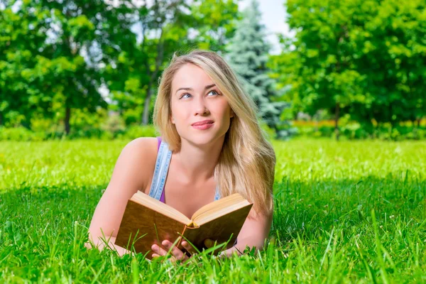 Mooie blonde liggen op het gras en bezit in handen dik boek — Stockfoto