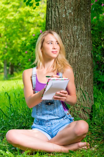 Blonde with a notebook and pen sitting near a tree — Stock Photo, Image