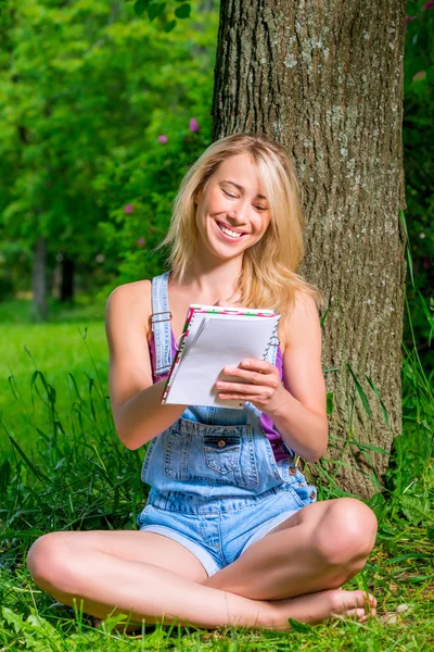 Glückliche blonde Frau mit Notizbuch und Stift, die neben einem Baum sitzt — Stockfoto