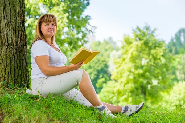 Beautiful woman 50 years with a book near a tree on the lawn in — Stock Photo, Image