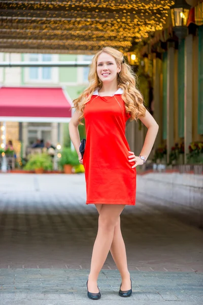Slender young girl in a red dress posing near the building — ストック写真