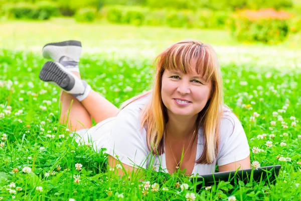 Mulher feliz deitado em um prado verde com um tablet em suas mãos — Fotografia de Stock