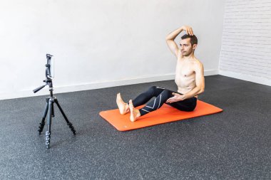 Paspasın üzerinde hipopresif abdominal egzersizler öğreten beyaz bir adam, öğrencileri için online ders veren tabletine bağlı.