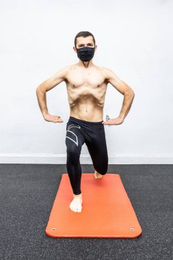 Yüzü maskeli beyaz bir adam paspasın üzerinde hipopresif abdominal egzersiz yapıyor.