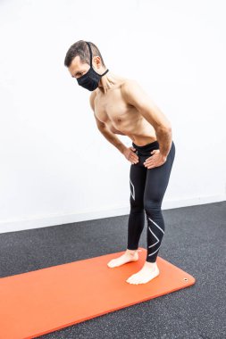 Yüzü maskeli beyaz bir adam paspasın üzerinde hipopresif abdominal egzersiz yapıyor.