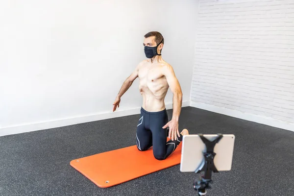 caucasian man with a face mask training hypopressive abdominal exercises on the mat, connected to his tablet performing online class for his students