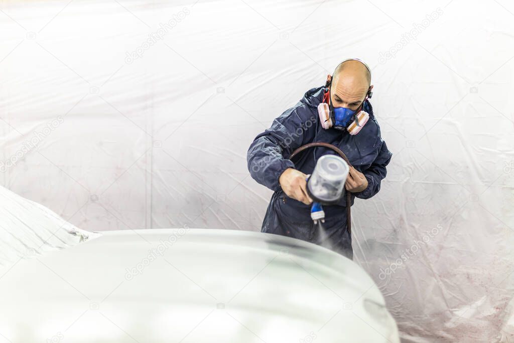 young adult male car painter, painting a car in the paint booth of his workshop