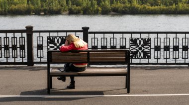 Krasnoyarsk, Rusya - 9 Eylül 2019: Krasnoyarsk yakınlarındaki Yenisej nehrinin kenarındaki bankta oturan kırmızı ceketli yalnız kız.