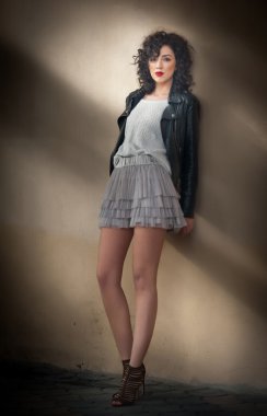 Sevimli genç kıvırcık esmer kadın dantel kısa etek ve siyah deri ceket bir duvara yaslanmış. Duvarının yakınında uzun bacaklı seksi güzel genç kadın. Şehvetli kız poz tam uzunlukta portresi