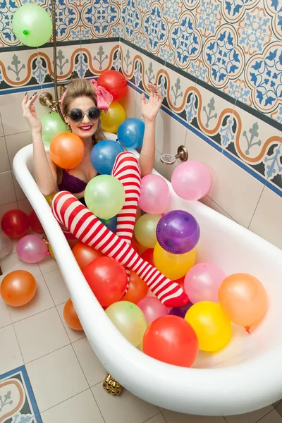 Blonde Frau mit Sonnenbrille spielt in ihrer Badewanne mit bunten Luftballons. sinnliche Mädchen mit weiß-rot gestreiften Strümpfen, die Spaß im Badezimmer haben, bedeckt mit Luftballons — Stockfoto