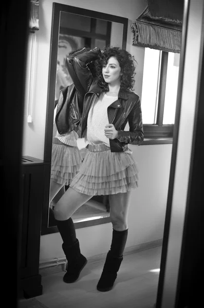 Young woman in black leather jacket and gray short tutu skirt looking into a large mirror. Beautiful curly dark hair girl posing in front of a wall mirror, indoors shot. Fashionable brunette model. — Stock Photo, Image