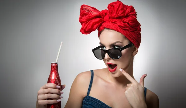 Kırmızı türbanlı ve güneş gözlüklü bir kadın elinde pipetle içki şişesi tutuyor. Elinde şişe olan çekici bir kız portresi, gri arka planda stüdyo resmi. Mutlu genç bayan, reklam konsepti. — Stok fotoğraf