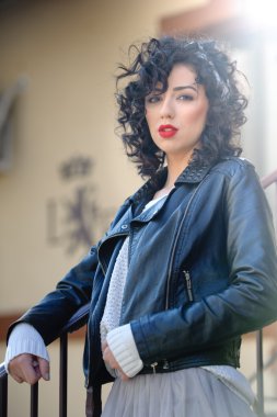 Charming young curly brunette woman with black leather jacket. Sexy gorgeous young woman with modern look. Portrait of sensual girl with voluptuous mouth wearing bright red lipstick, outdoor shot