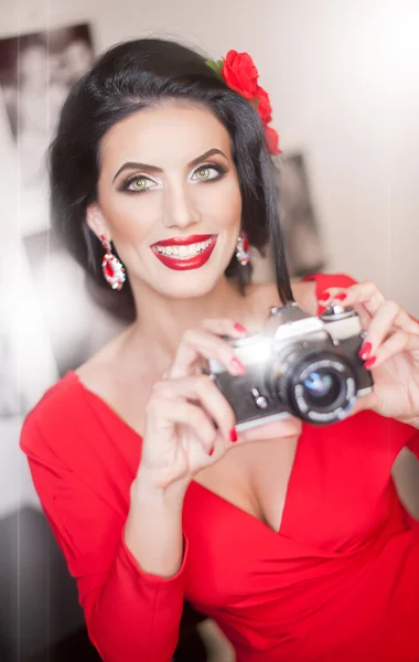 Hermosa mujer joven con maquillaje creativo y estilo de pelo tomando fotos con una cámara. Morena atractiva de moda con look español sosteniendo una cámara. Dama de rojo con flor en el pelo, sonriendo — Foto de Stock