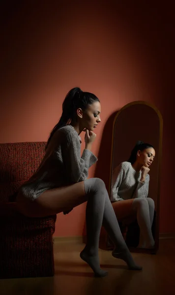 Attractive sexy brunette in white comfortable pullover posing sitting on a sofa indoors. Portrait of sensual young woman posing in front of large mirror. Beautiful girl with long stockings in her room — Stock Photo, Image