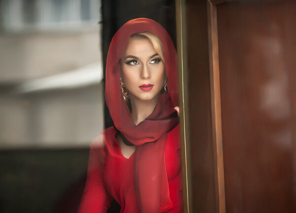 Charming young blonde with red blouse and headscarf posing in door frame. Sensual gorgeous young woman in red outfit opening the door for somebody.Portrait of attractive lady in red with romantic look