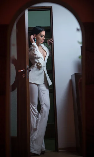 Joven mujer morena hermosa en elegante traje blanco con pantalones de pie en el marco de la puerta. Chica seductora de cabello oscuro posando en interiores. Elegante dama en traje blanco con chaqueta con escote hundido — Foto de Stock