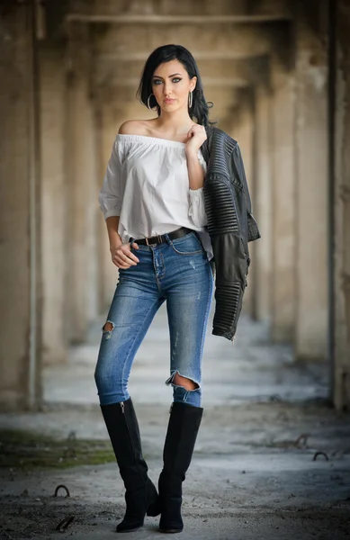 Retrato de una hermosa joven sexy con atuendo moderno, chaqueta de cuero, jeans, blusa blanca y botas negras, en el fondo urbano. Atractiva joven morena con el pelo largo y ojos azules posando . —  Fotos de Stock