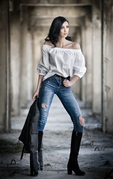 Retrato de una hermosa joven sexy con atuendo moderno, chaqueta de cuero, jeans, blusa blanca y botas negras, en el fondo urbano. Atractiva joven morena con el pelo largo y ojos azules posando . —  Fotos de Stock