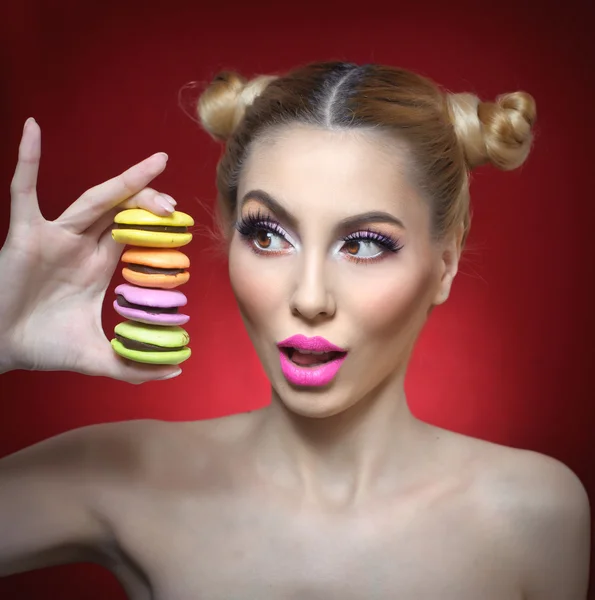 Beautiful model with makeup and creative hairstyle holding colorful macaroons, studio shoot on red background. Diet, dieting concept. Sweets. Sexy woman with pink lipstick showing a pile of macaroons — Stock Photo, Image