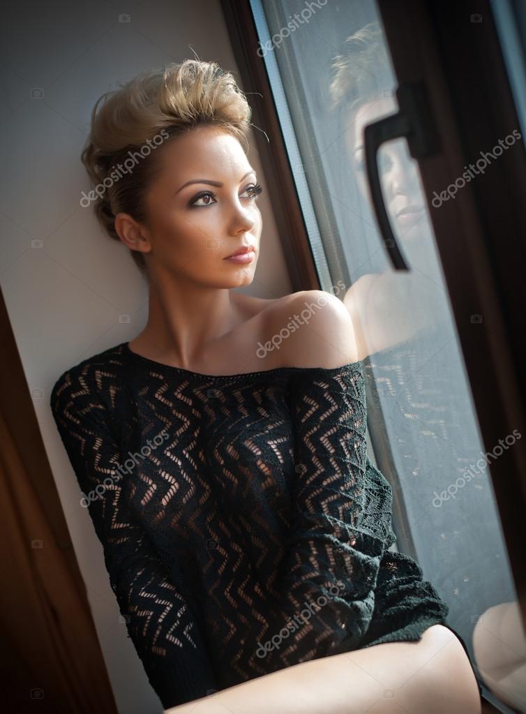 Voorkomen Krijgsgevangene Feest Attractive sexy blonde with black see through blouse looking on the window  in daylight. Portrait of sensual short fair hair woman wearing low shoulder  top, indoor scene. Beautiful woman daydreaming Stock Photo