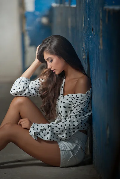 Beautiful girl with unbuttoned shirt posing, old wall with peeling blue paint on background. Pretty brunette sitting on the floor. Attractive dark long hair young woman with sensual expression on face — Stock Photo, Image