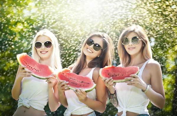 Hermosas chicas con gafas de sol comiendo sandía fresca y sonriendo. Mujeres jóvenes felices comiendo sandía en el parque. Estilo de vida juvenil. Felicidad, alegría, concepto de verano . — Foto de Stock
