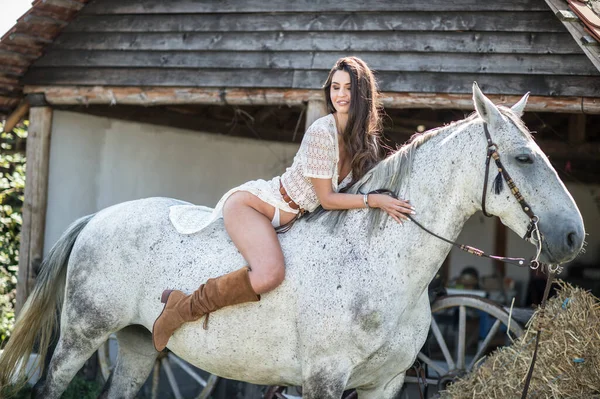 長い髪と白い馬に乗って長い白いドレスを持つ美しいブルネットの女性 官能的で魅力的な女の子で茶色のブーツが相互作用し 牧場で馬と楽しむ — ストック写真