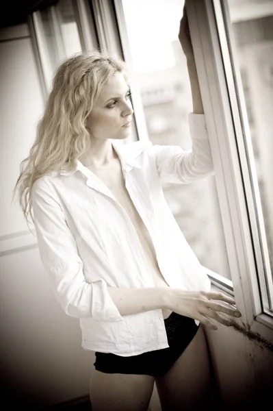 Attractive sexy blonde with white shirt looking on the window in daylight. Portrait of sensual long fair hair woman wearing blouse and black panties indoor scene. Beautiful long hair woman daydreaming — Stock Photo, Image