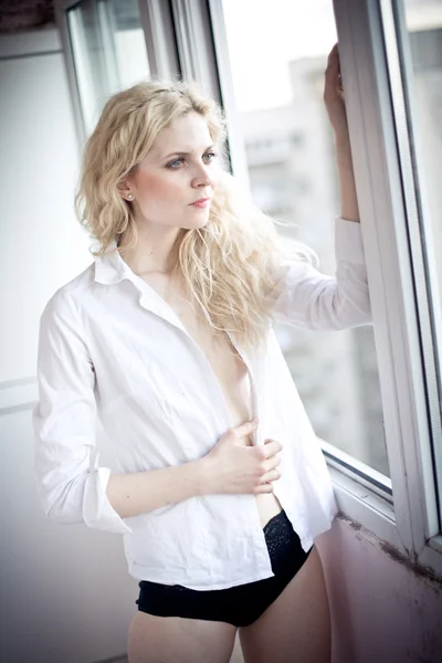 Attractive sexy blonde with white shirt looking on the window in daylight. Portrait of sensual long fair hair woman wearing blouse and black panties indoor scene. Beautiful long hair woman daydreaming — Stock Photo, Image