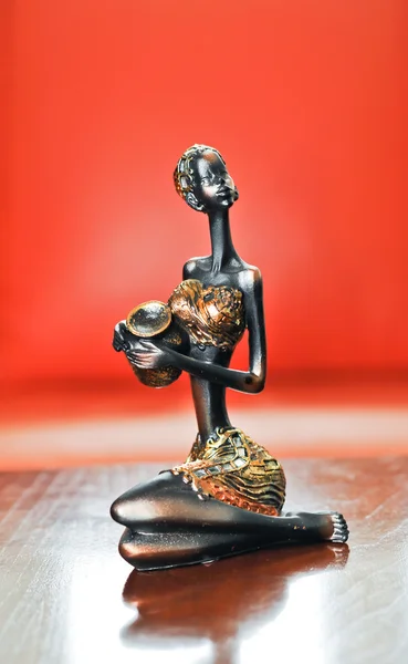 Statua di donna africana seduta e con in mano una pentola. Miniatura d'oro di giovani donne su sfondo rosso . — Foto Stock