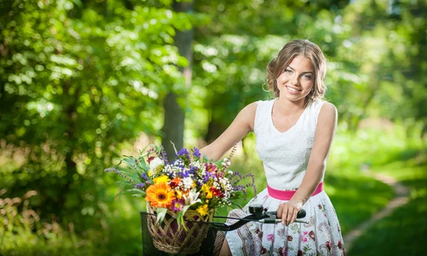 Bella ragazza che indossa un bel vestito bianco divertirsi nel parco con la bicicletta. Sano concetto di stile di vita all'aperto. Paesaggio d'epoca. Bella ragazza bionda con look retrò con bici e cesto con fiori — Foto Stock