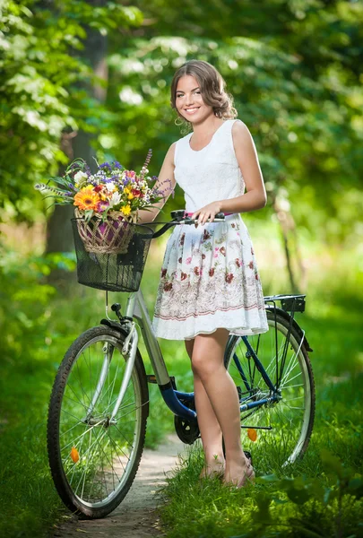 Menina bonita vestindo um vestido branco agradável se divertindo no parque com bicicleta. Conceito de estilo de vida saudável ao ar livre. Cenário vintage. Menina loira bonita com olhar retro com bicicleta e cesta com flores — Fotografia de Stock