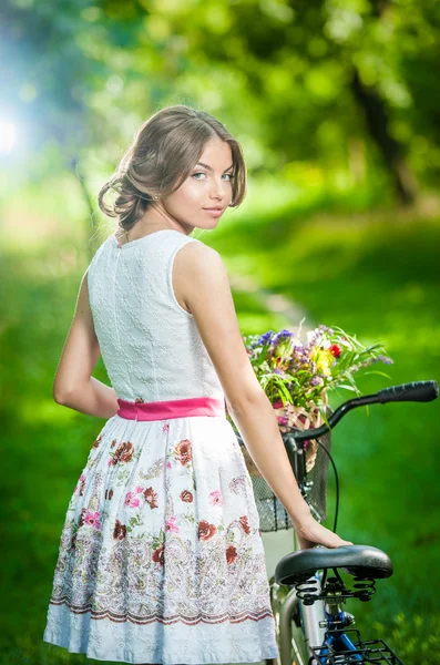 Menina bonita vestindo um vestido branco agradável se divertindo no parque com bicicleta. Conceito de estilo de vida saudável ao ar livre. Cenário vintage. Menina loira bonita com olhar retro com bicicleta e cesta com flores — Fotografia de Stock